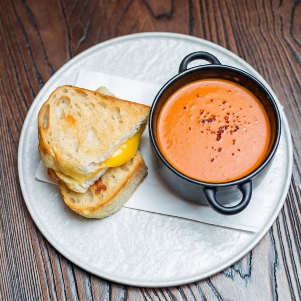 Grilled cheese sandwich with melted yellow cheese and a black bowl of tomato soup on a white plate.