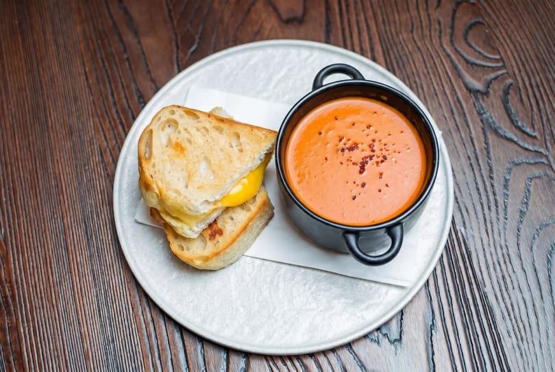 Grilled cheese sandwich with melted yellow cheese and a black bowl of tomato soup on a white plate.