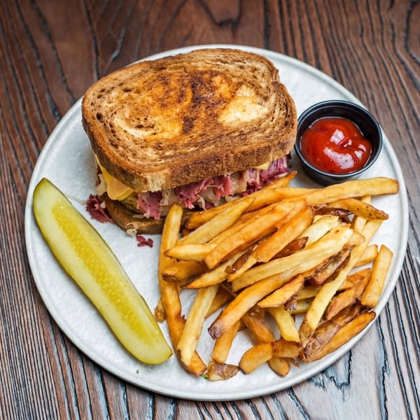 Reuben sandwich with fries, a pickle spear, and ketchup on a white plate on a wooden table.