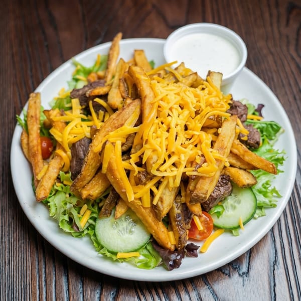 Steak salad with french fries, shredded cheddar cheese, and a side of ranch dressing.