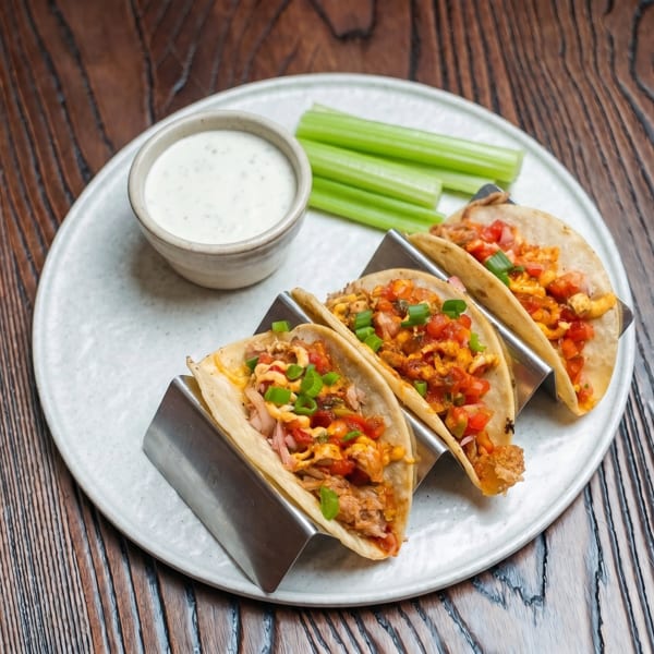 Three tacos in metal holders with salsa, green onions, and a side of ranch and celery sticks.