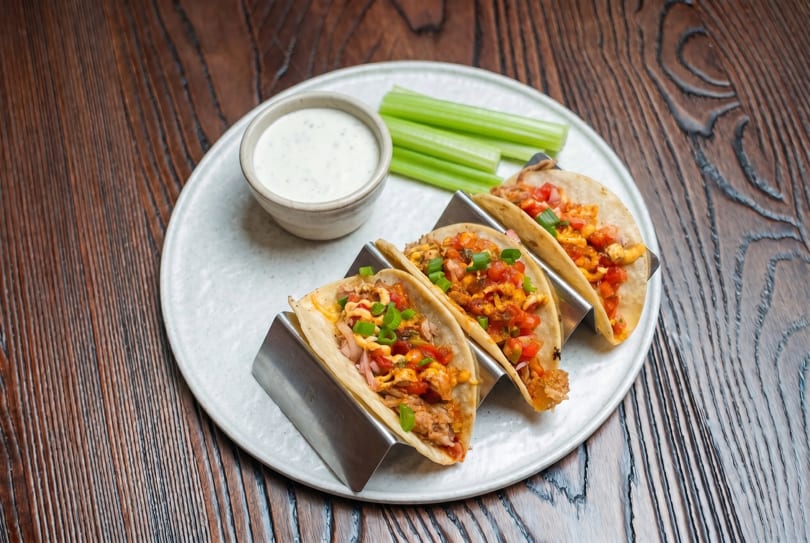 Three tacos in metal holders with salsa, green onions, and a side of ranch and celery sticks.